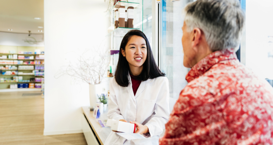 A female-presenting pharmacist explains a prescription to a white, male-presenting person in a patterned shirt while seated in a pharmacy setting.