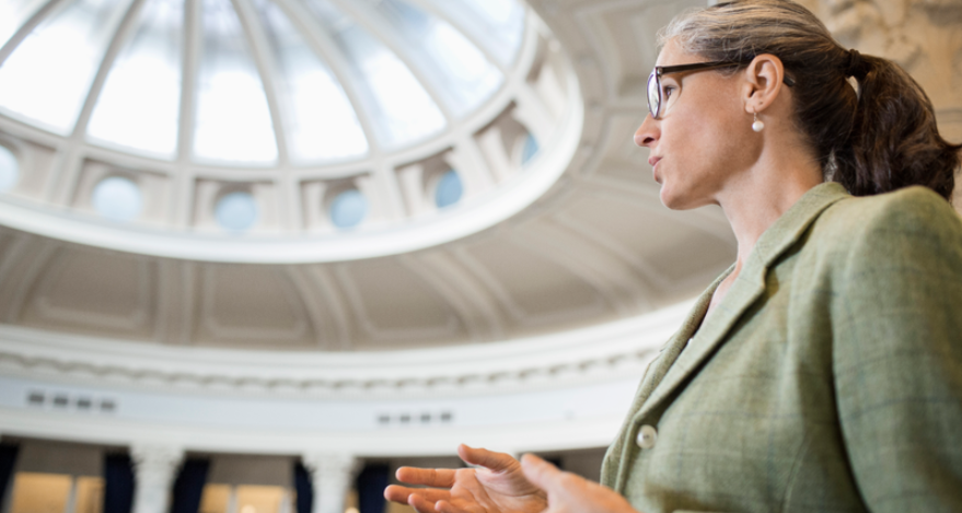 A female-presenting person speaks at a legislative building, wearing a green blazer and glasses.