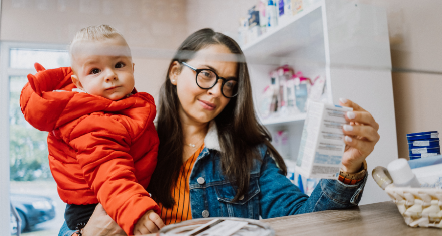 A woman holds up a prescription medication at her pharmacy while holding a small child in her other hand.
