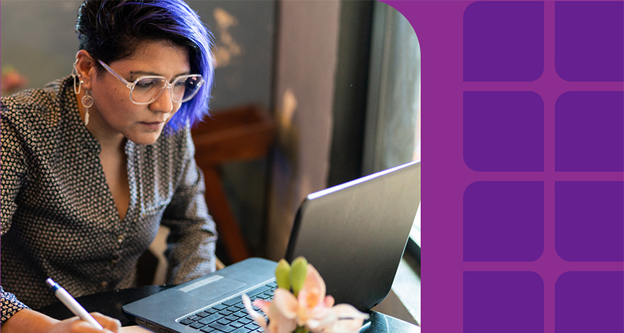 A person with purple hair and glasses works at a laptop with a pen in their hand