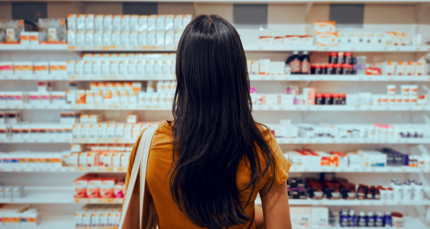 A woman with long, dark hair looks at a shelf full of hundreds of medications at a pharmacy.