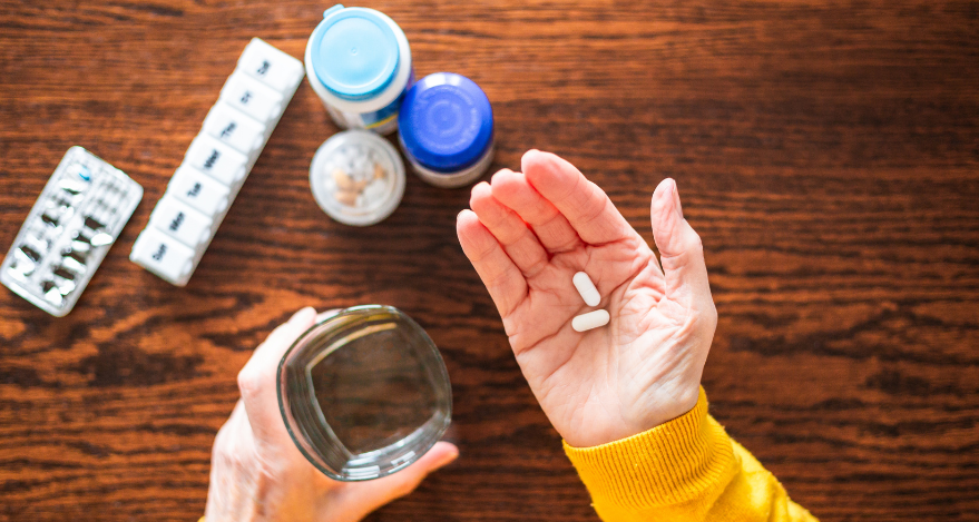 A person holds a glass of water in their left hand and a couple of pills in their right hand. In the background, you can see a table with a pill box and a few medication bottles.