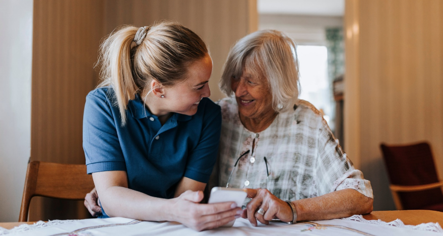 An older woman and a younger woman smile at each other while using a smartphone.