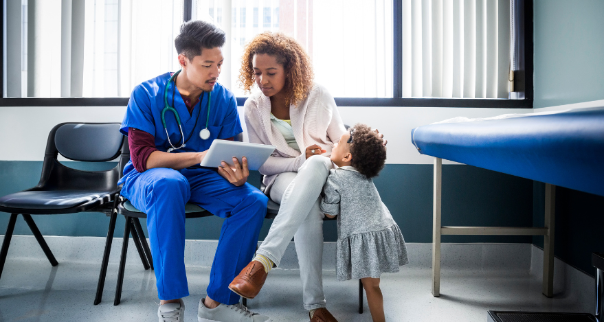A woman sits next to a doctor while a young child pulls at her sweater.