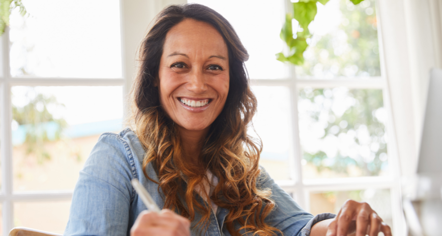 A woman sits at a desk and writes something with a pen in her hand. She smiles at the camera.