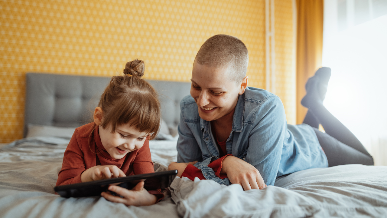 A woman with extremely short hair lies down next to a toddler on a tablet. Both individuals are smiling.